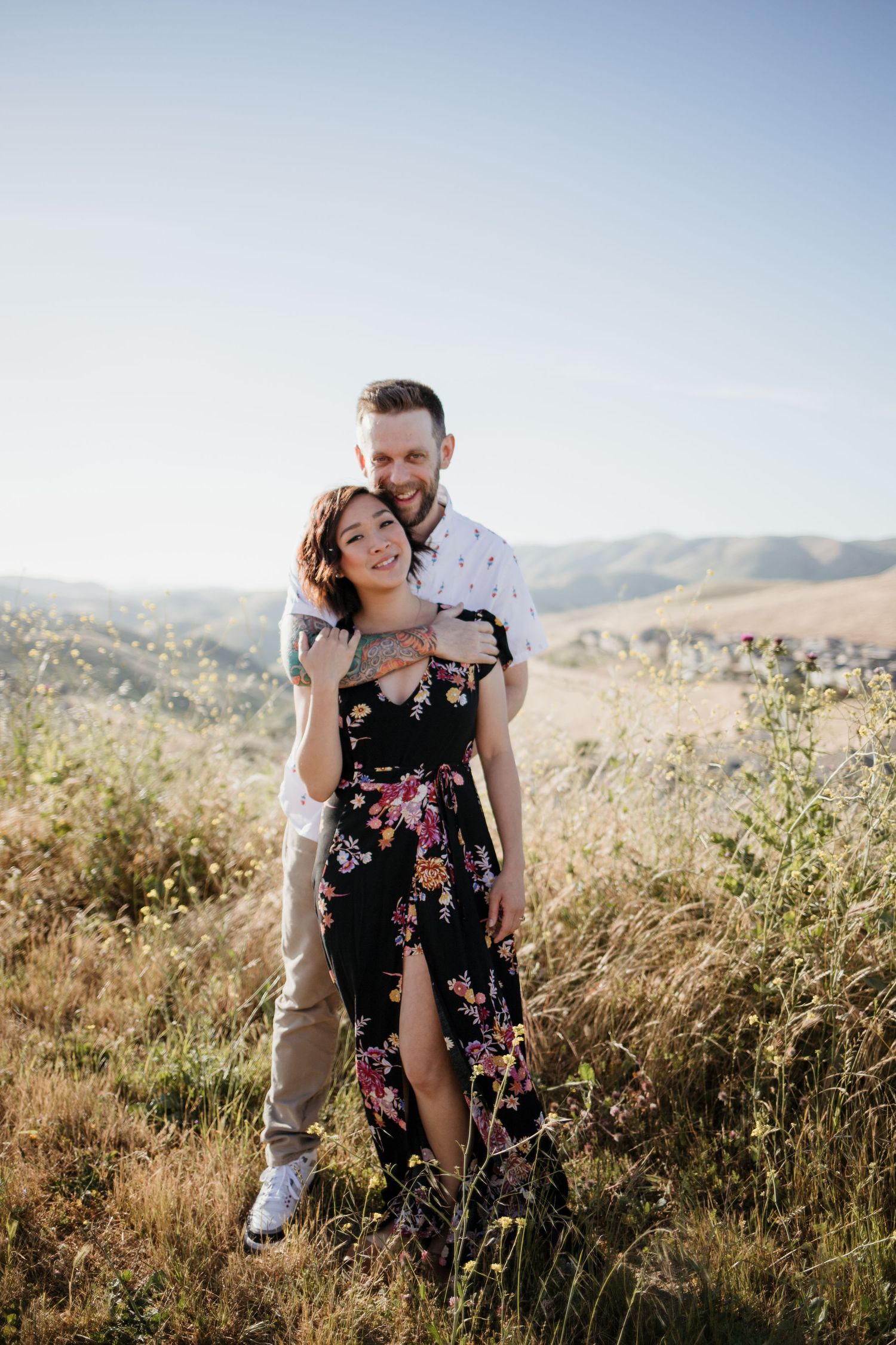 Couple embracing in a sunny meadow, woman in floral dress, rolling hills in background.