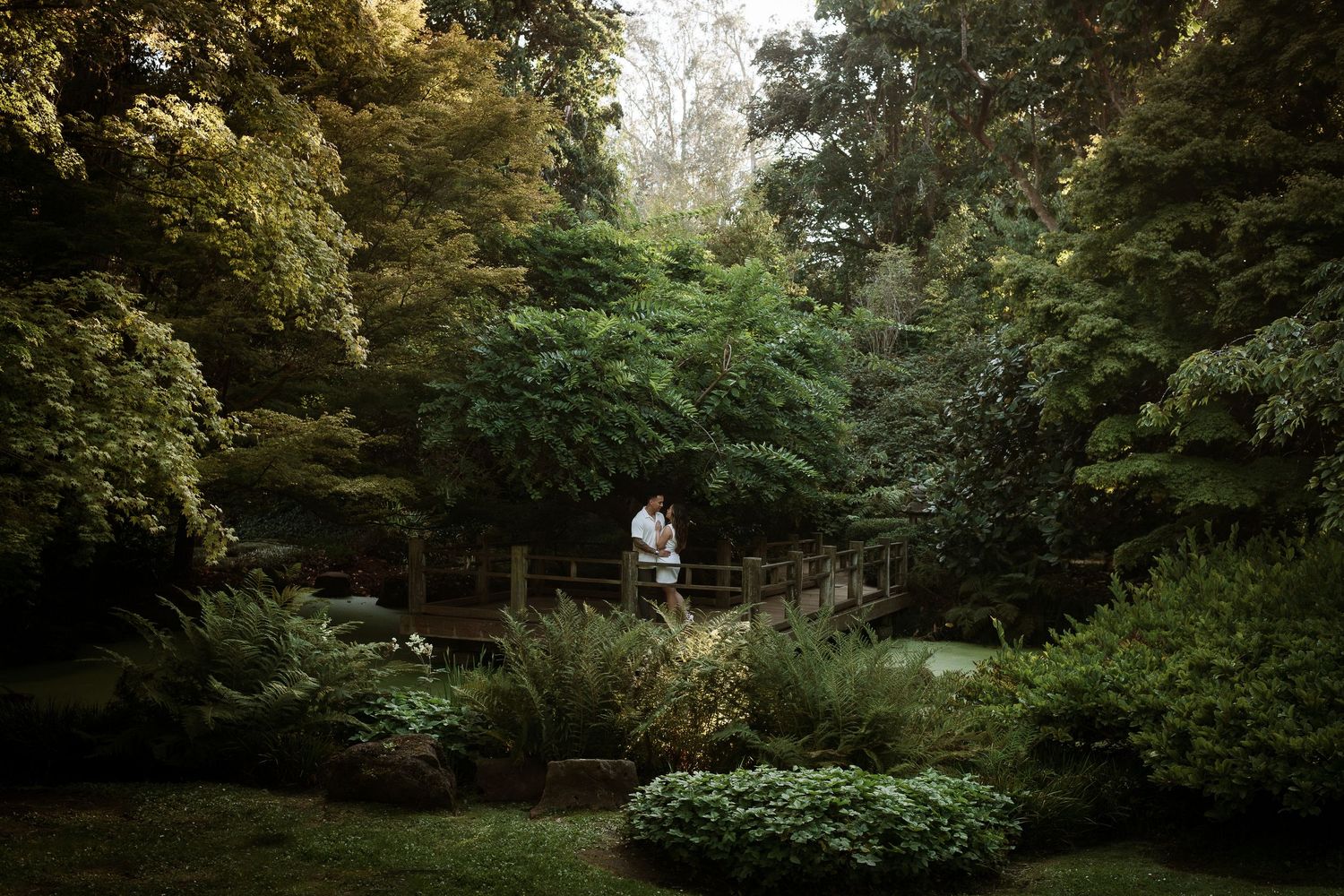 Couple embracing on a wooden bridge surrounded by lush green trees in a serene garden setting.