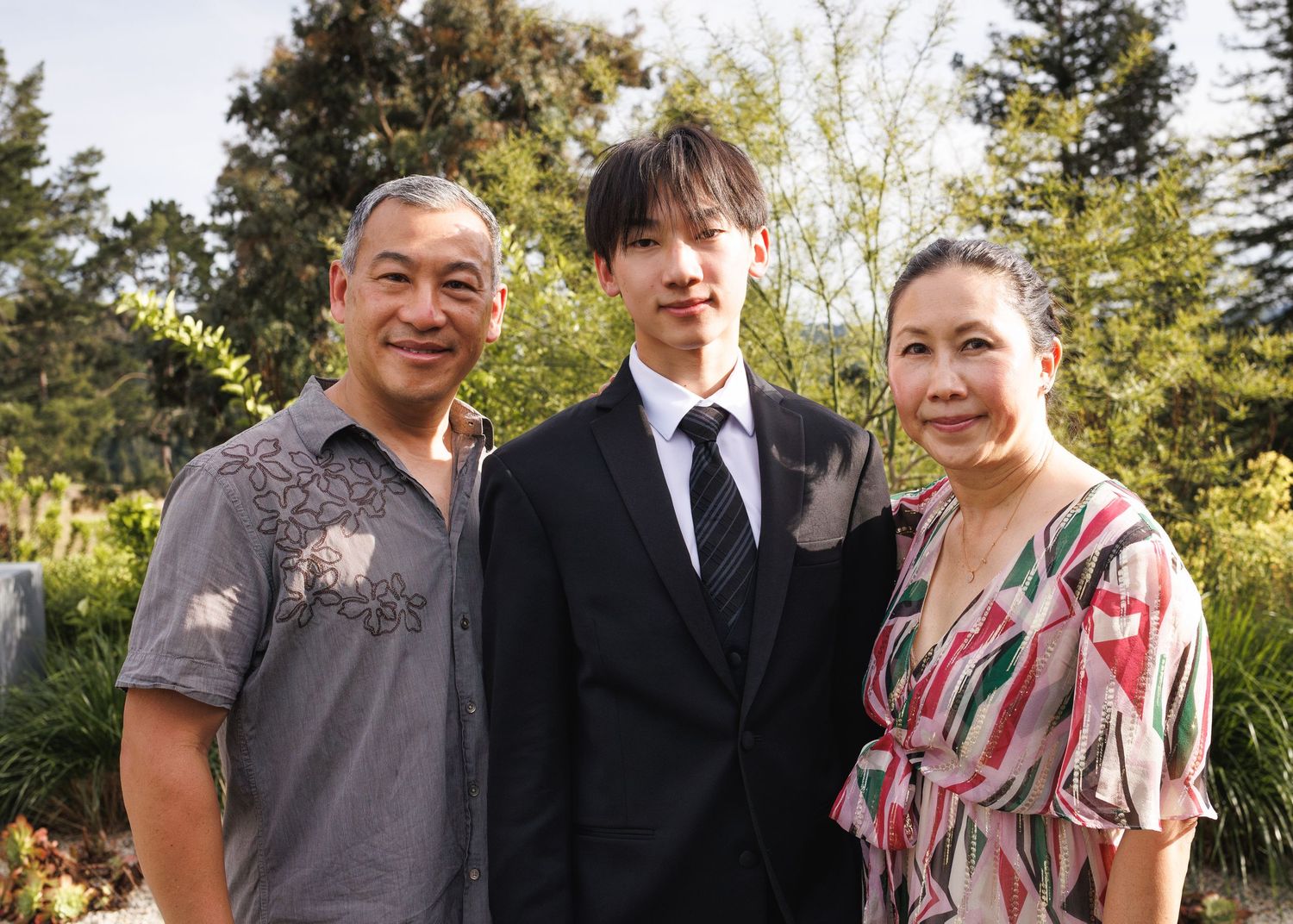 Family of three posing outdoors in a garden setting, dressed in formal and casual attire for a special occasion.