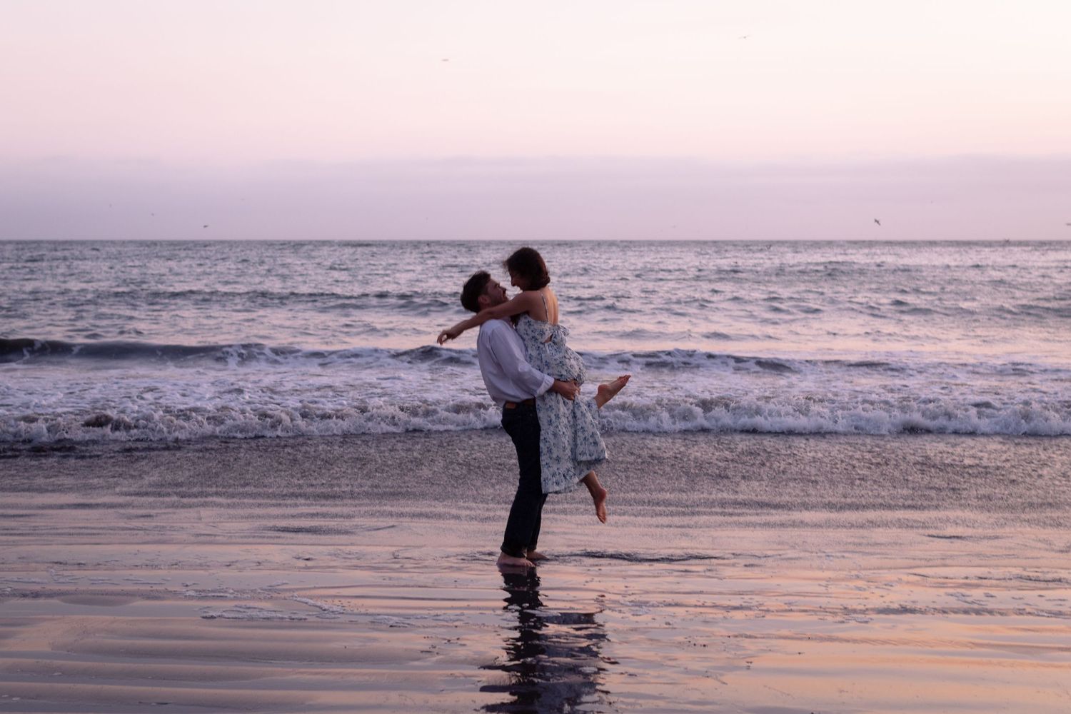 Couple embracing romantically on a sandy beach at sunset with ocean waves in the background.