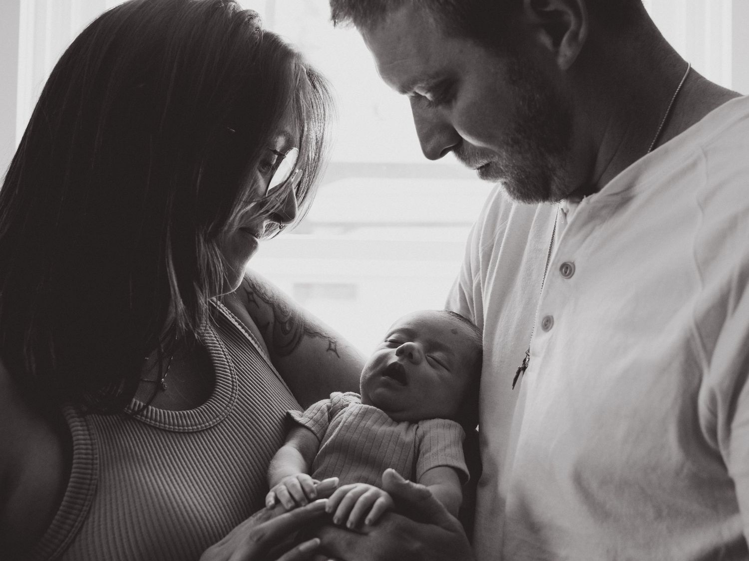 Black and white photo of new parents lovingly gazing at their sleeping newborn baby together.