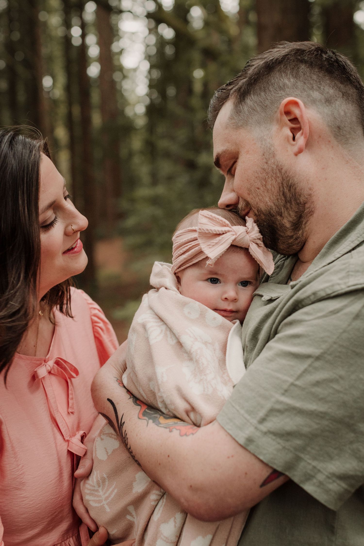 A couple in a redwood forest tenderly holding their newborn baby girl wearing a pink bow headband in Oakland, California.