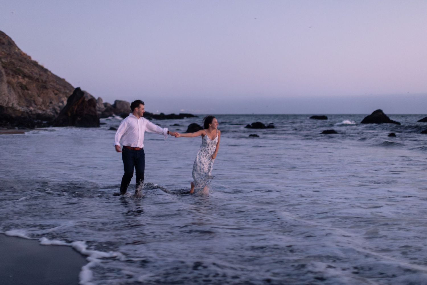 Couple holding hands walking through ocean waves at sunset on a rocky beach in a romantic engagement photo.
