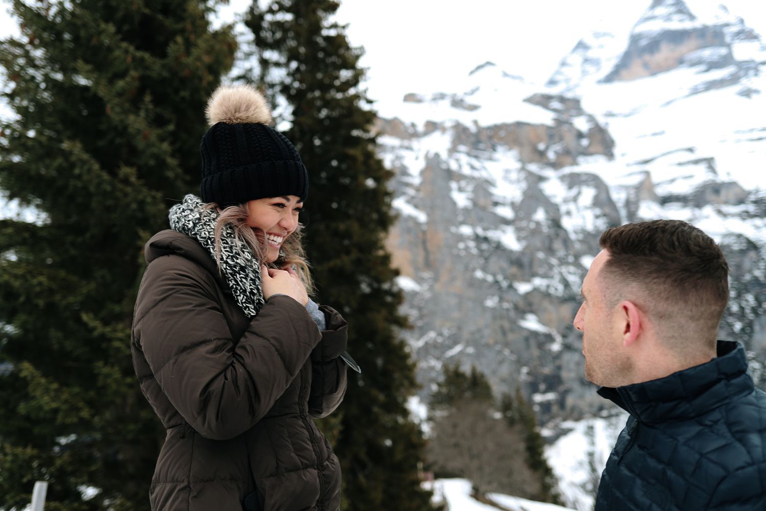 Woman in black pom-pom hat reacts emotionally during a snowy mountain proposal scene.