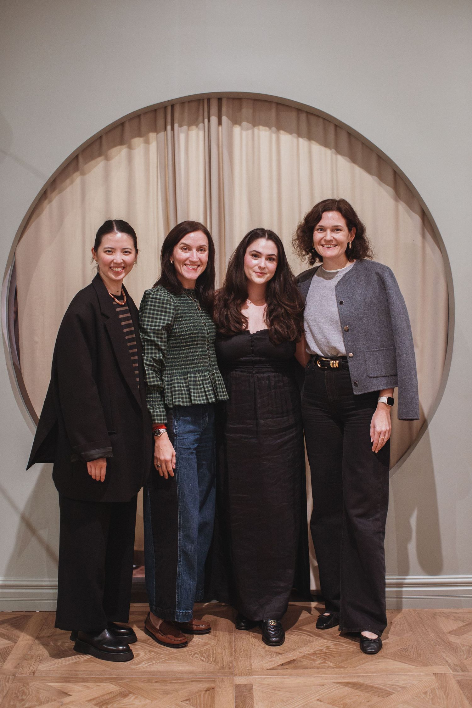 Four women smiling together in front of a circular mirror backdrop in an elegant interior setting.