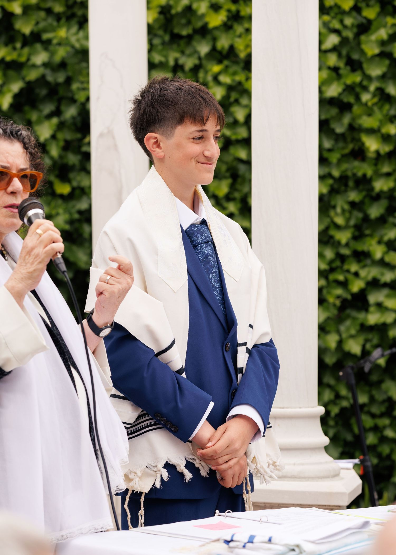 Person in blue suit and white tallit smiles during an outdoor Jewish ceremony beside a speaker with microphone.