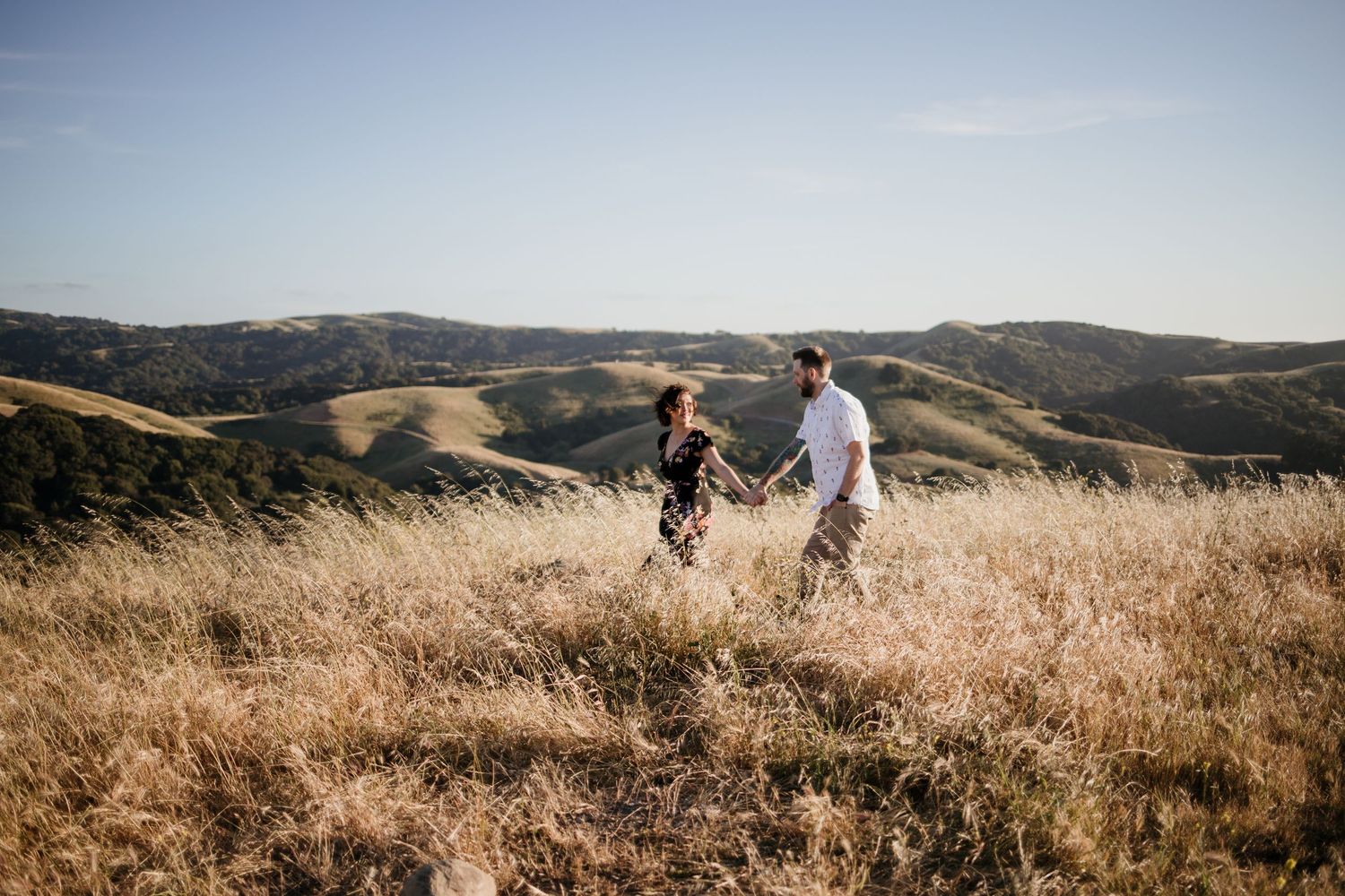 Couple holding hands walking through golden grass fields with rolling hills and blue sky backdrop.