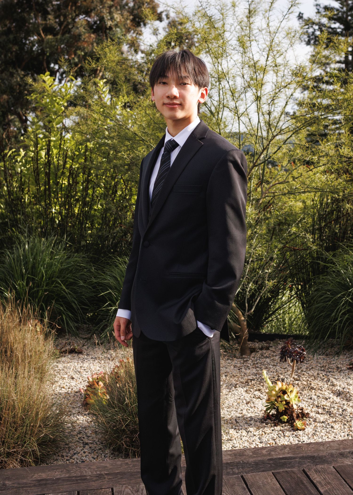 Young man in black suit and tie posing outdoors in a garden setting with lush green trees in background.
