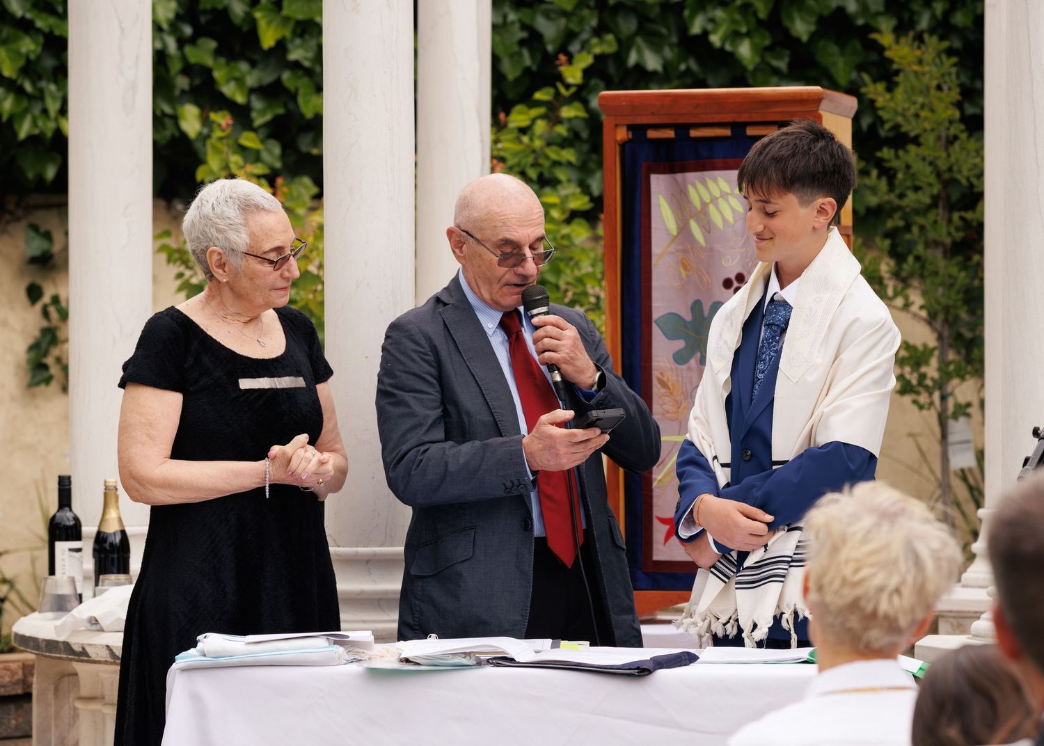 Three people stand outdoors at a formal gathering, with an older couple and a young man holding a microphone.