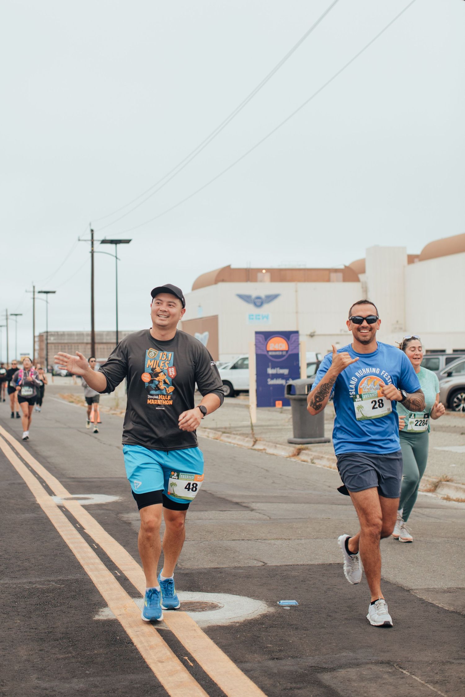 Two smiling runners wearing race bibs compete in an outdoor road race past a building on a cloudy day.
