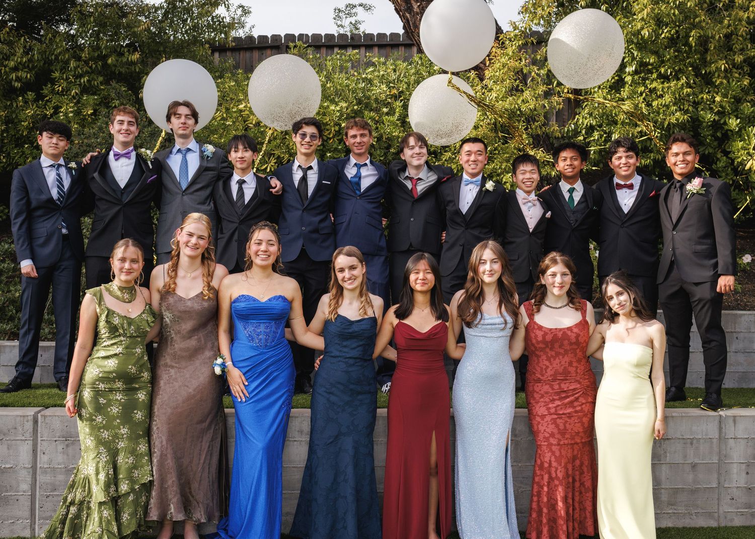 Large group of high school students pose outdoors in formal prom attire with white balloon decorations behind them.