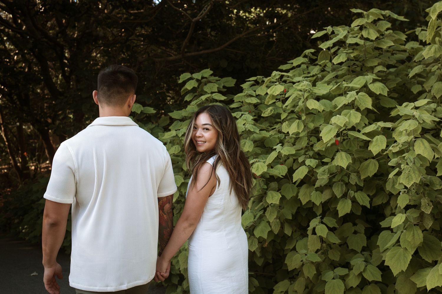 Couple in white outfits holding hands near lush green foliage, woman smiling back over shoulder outdoors.