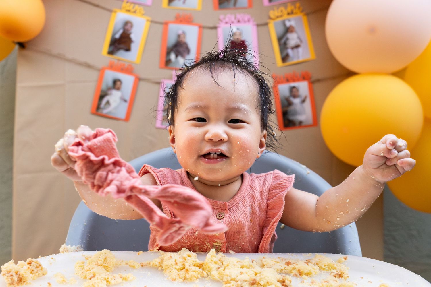 Happy baby girl celebrating first birthday smashing cake in high chair with balloons and photo banner.