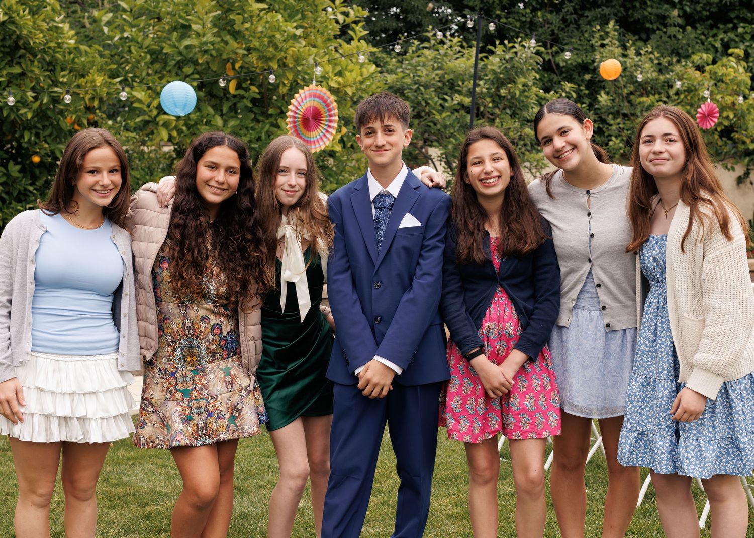Group of seven teenagers posing together outdoors at a garden party, dressed in formal attire with colorful decorations.