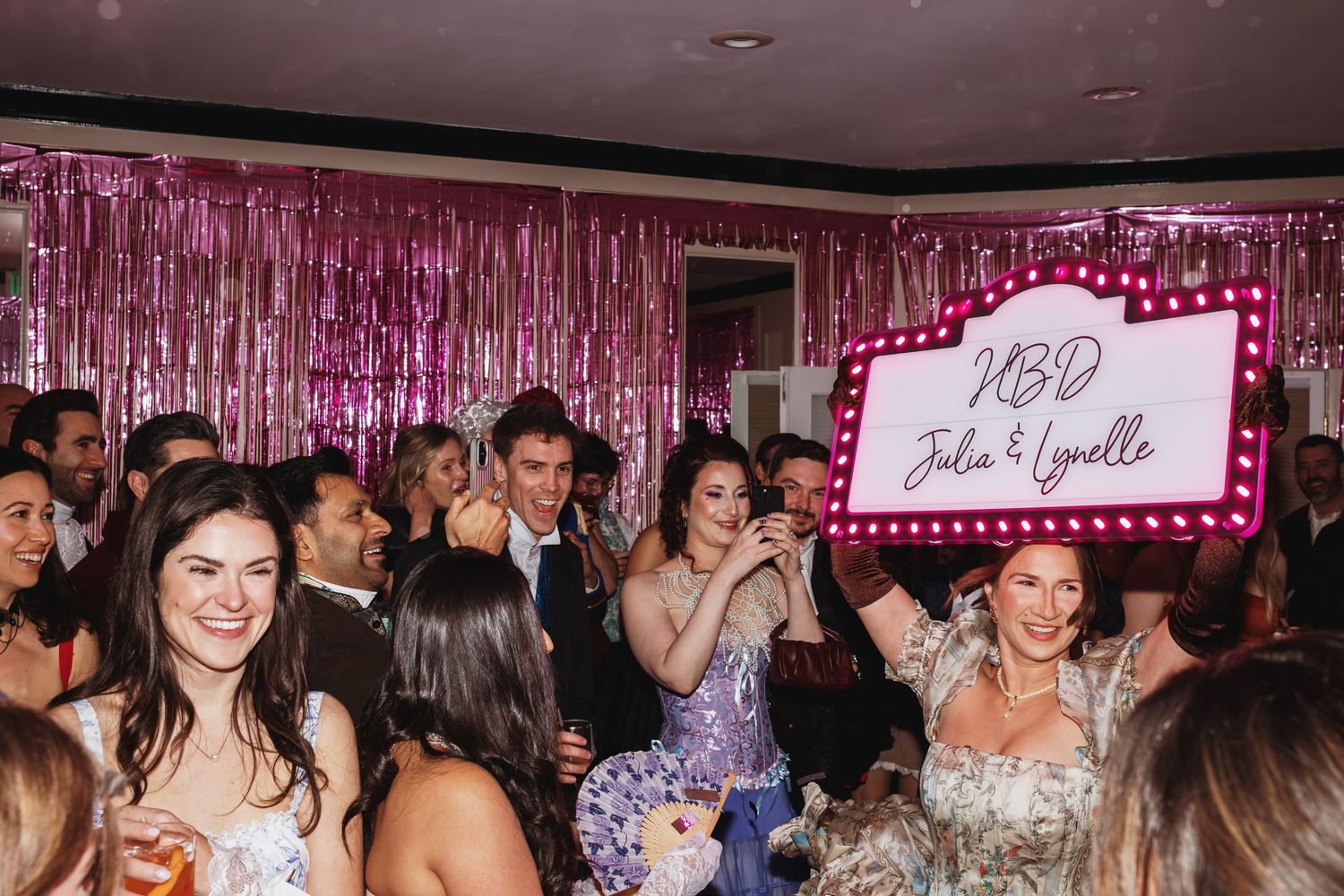 Guests celebrate at a wedding reception with pink tinsel backdrop and illuminated HBD Julia and Lynelle marquee sign.