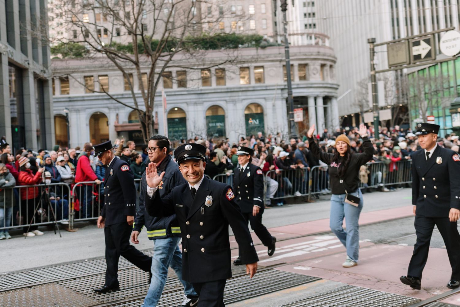 Firefighters in uniform march in a city parade along a crowded street lined with spectators behind metal barriers.