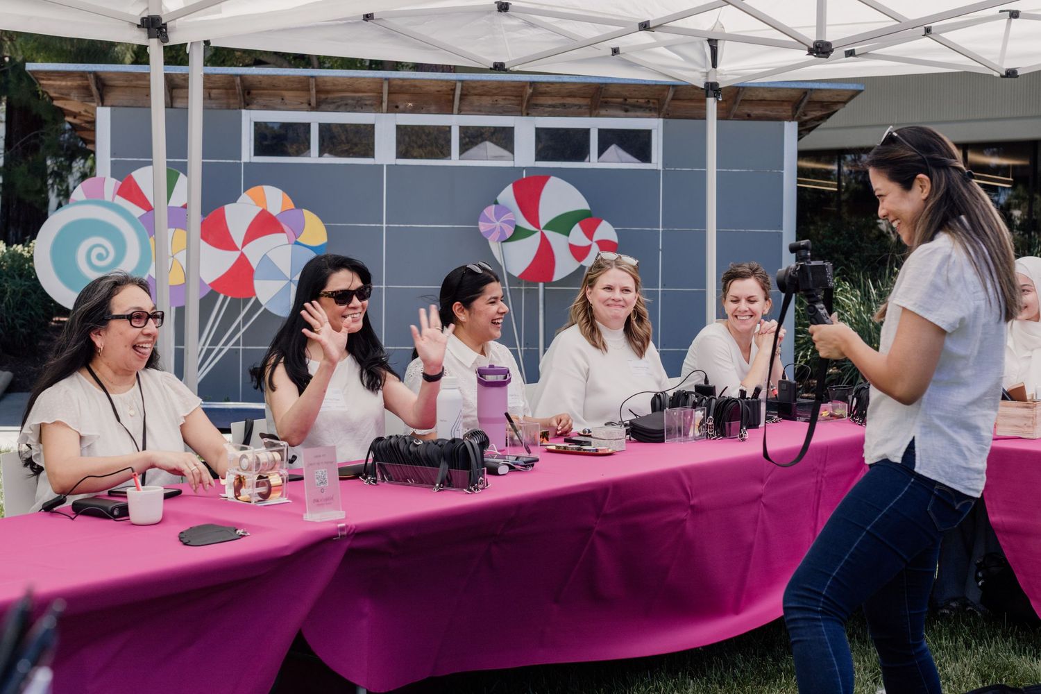 Women enjoy an outdoor makeup tutorial event at a pink table under a canopy with colorful candy decorations.