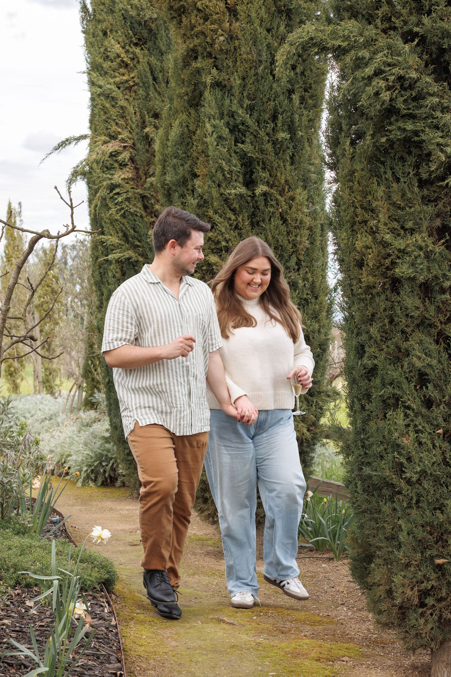 Couple walks hand-in-hand along a garden path lined with tall cypress trees, smiling at each other.