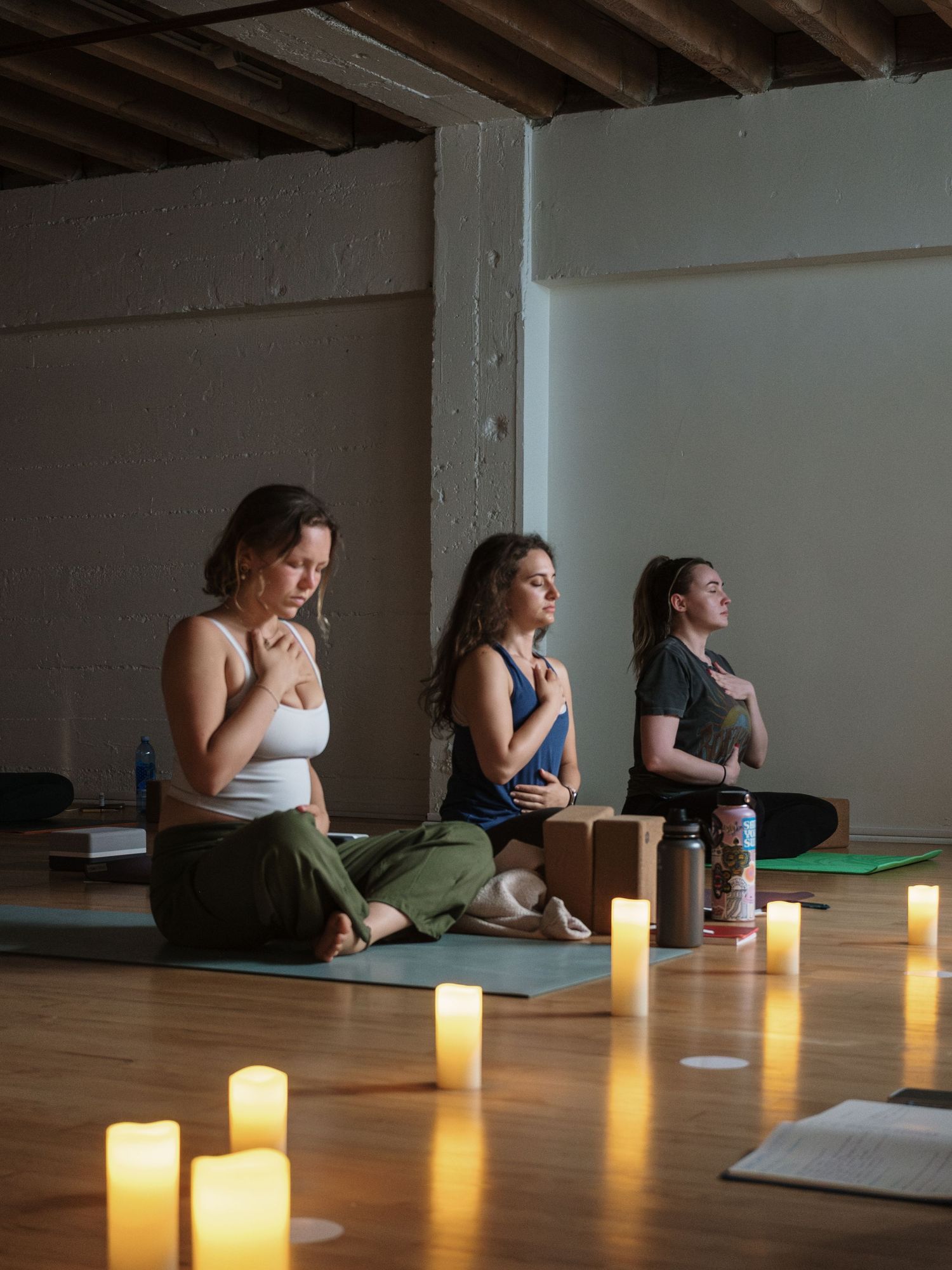 Three women sit cross-legged on yoga mats in candlelit studio, hands on hearts, eyes closed in meditative pose.