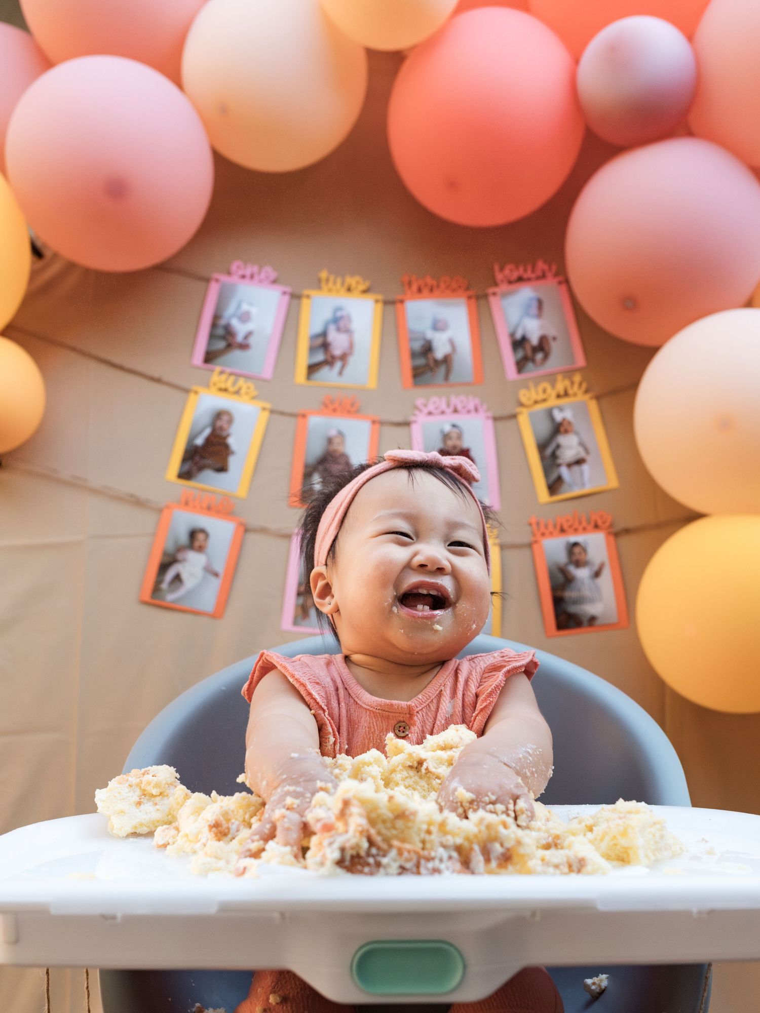 Baby girl laughing joyfully at her first birthday cake smash, surrounded by pink and peach balloons and monthly photo banner.