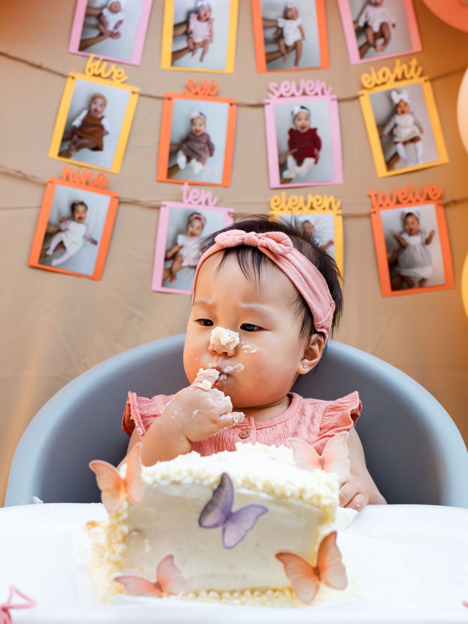 Baby girl in pink celebrates first birthday smashing white butterfly cake in high chair with monthly photo banner.