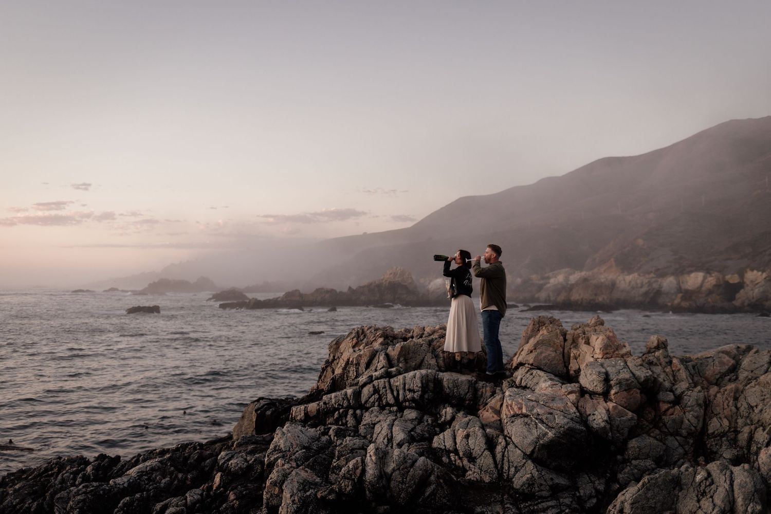 Couple standing on rocky coastal cliffs at sunset with misty mountains and ocean waves in the background.