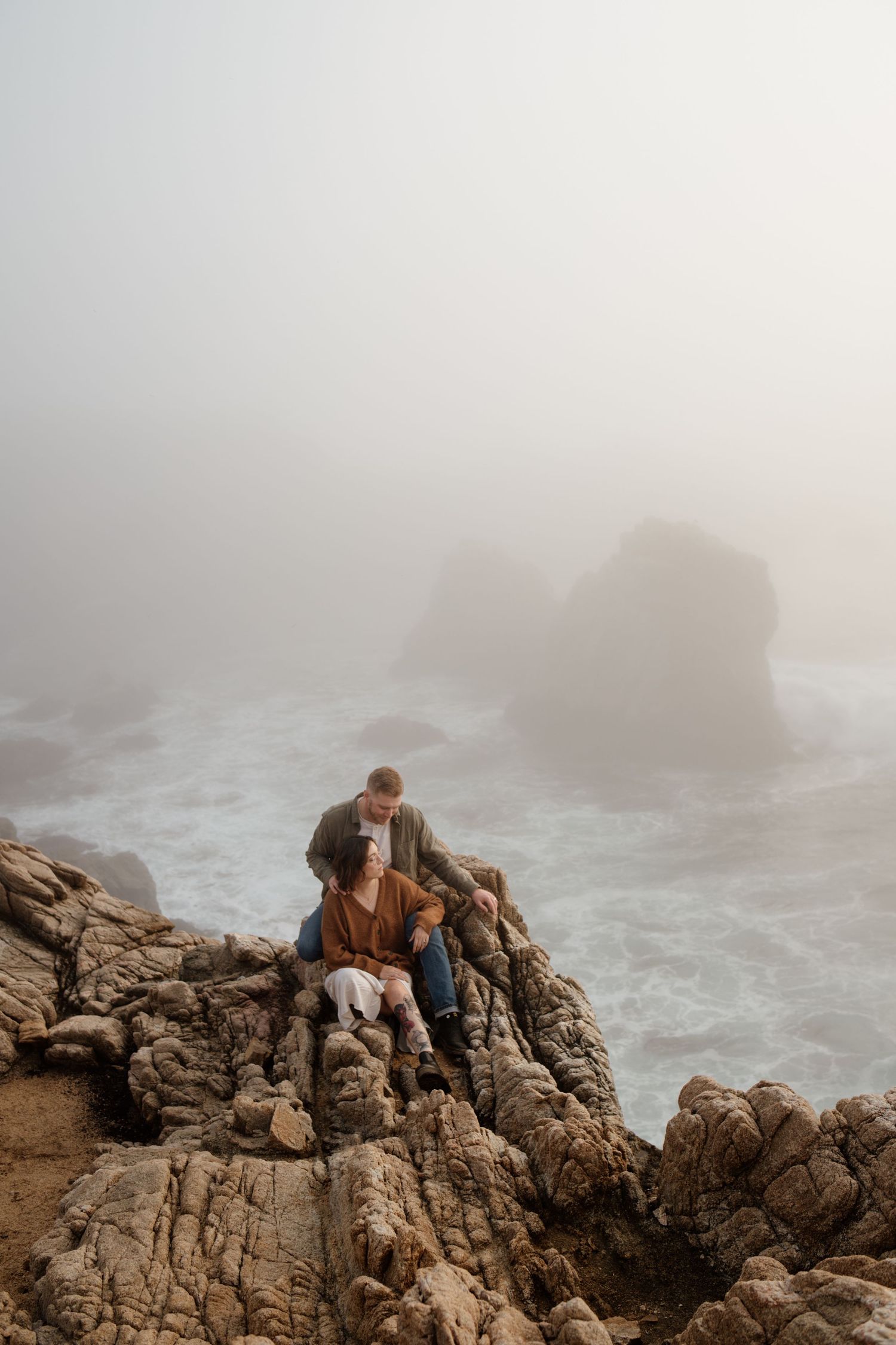 Couple sitting on rocky coastal cliffs in foggy weather, with misty ocean waves and sea stacks in the background.