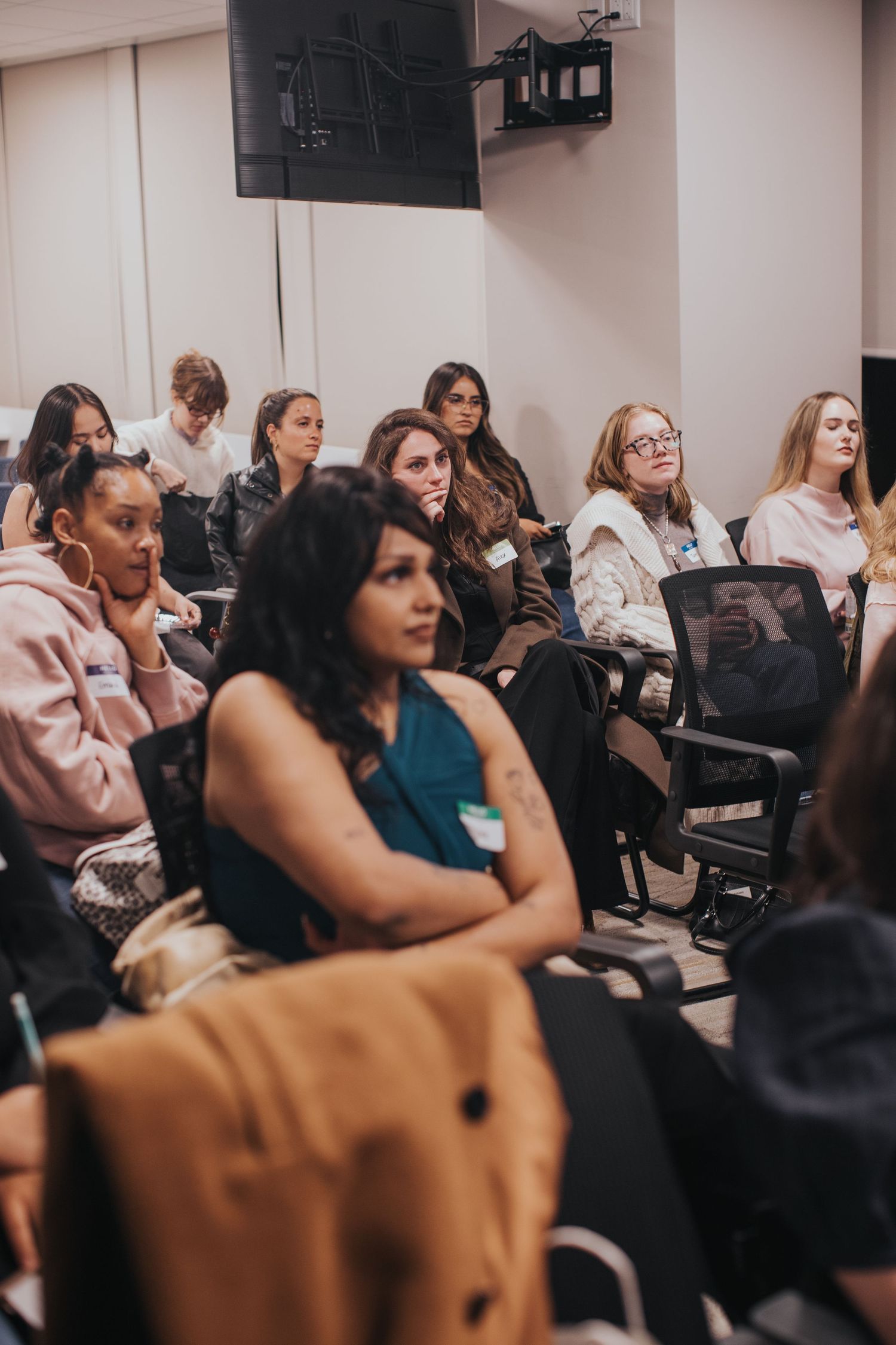 Diverse group of women seated in rows at an indoor seminar or conference, attentively listening to a presentation.