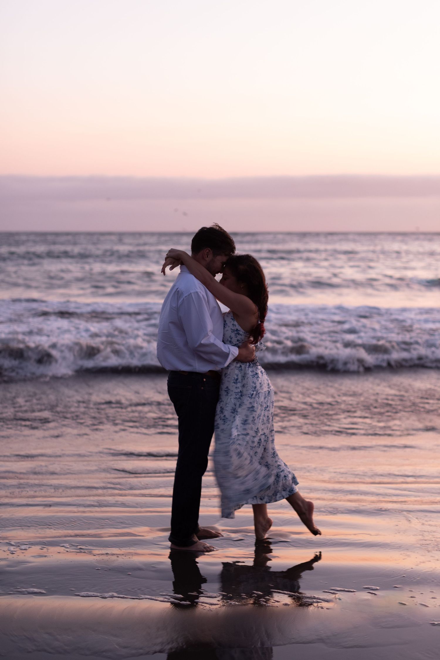 Couple embracing romantically on a wet beach at sunset, waves crashing behind them in soft pink light.
