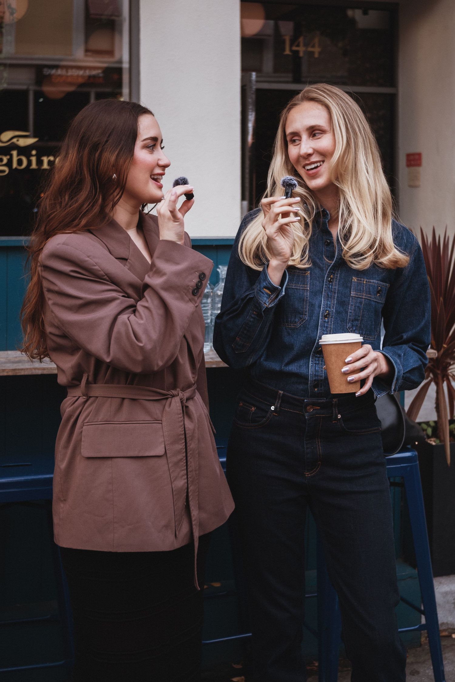 Two women smiling outdoors, one in a brown jacket and one in a denim shirt holding a coffee cup.