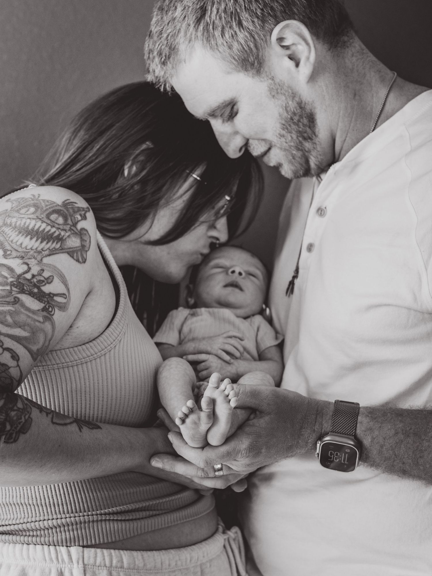 Black and white photo of parents lovingly gazing at their newborn baby, mother with tattoos, father wearing smartwatch.