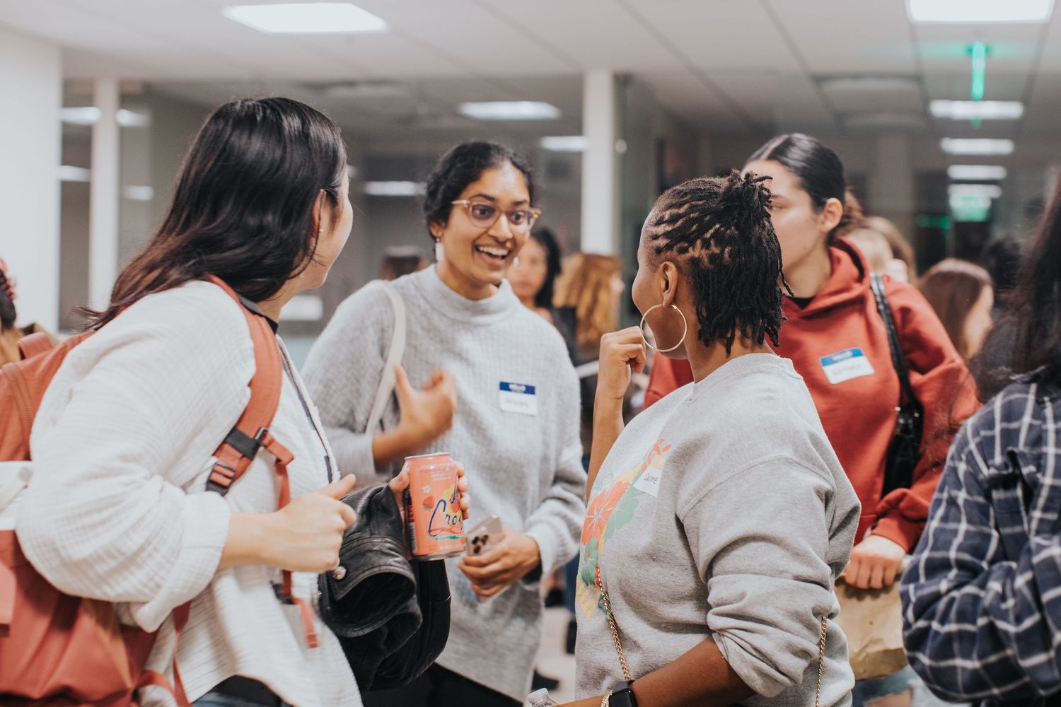 Diverse group of young women networking and conversing at an indoor college event wearing name tags.