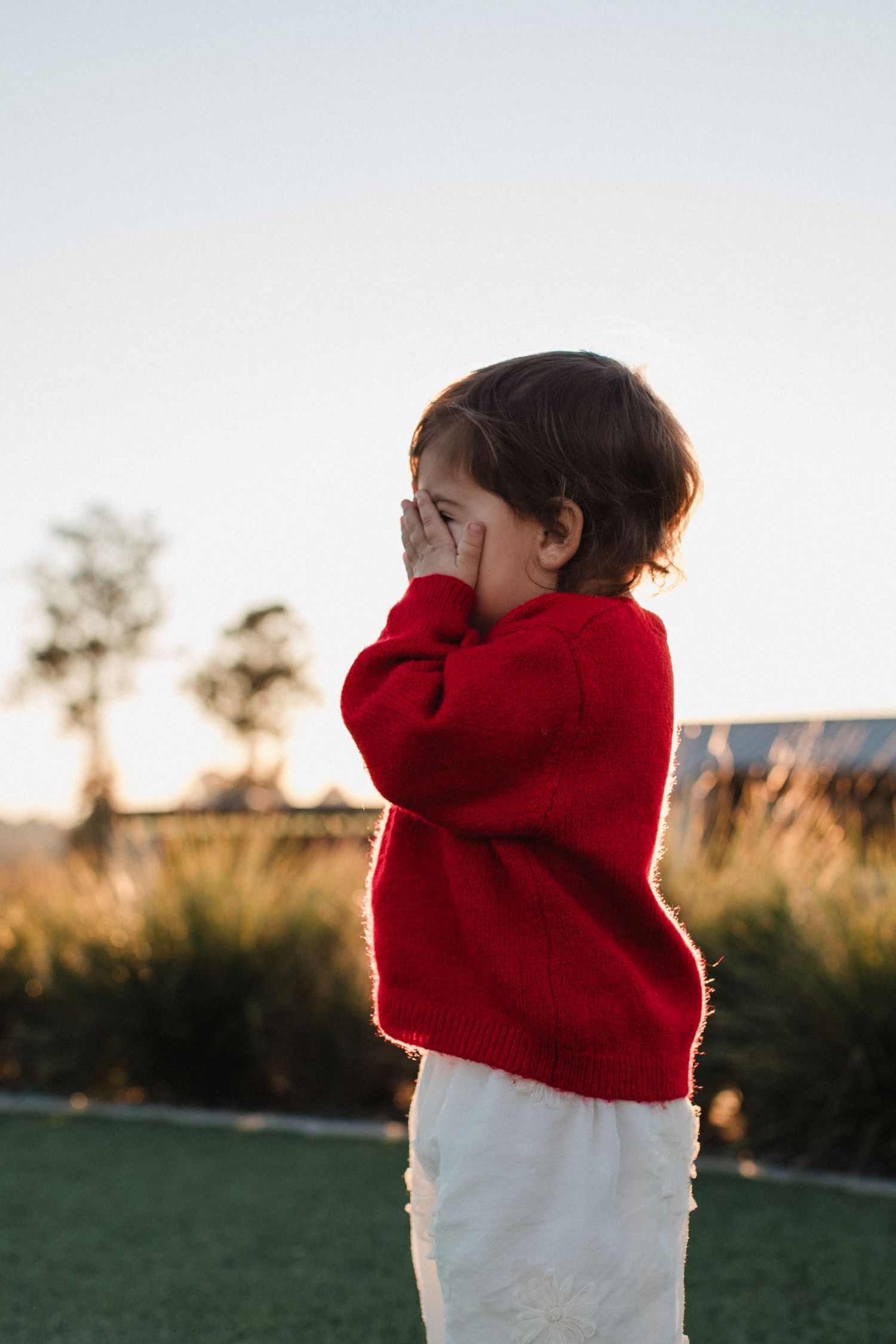 A child in a red hoodie plays peek-a-boo in a grassy field during sunset.