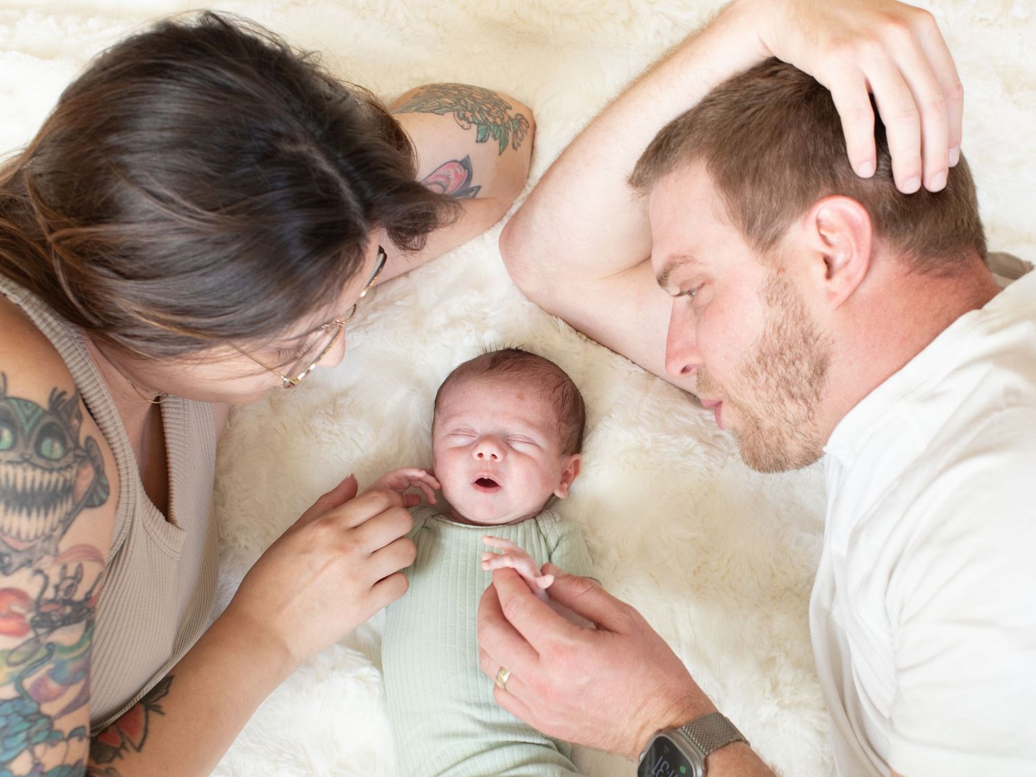 New parents lying beside their swaddled newborn baby on a soft white blanket, sharing a tender family moment.