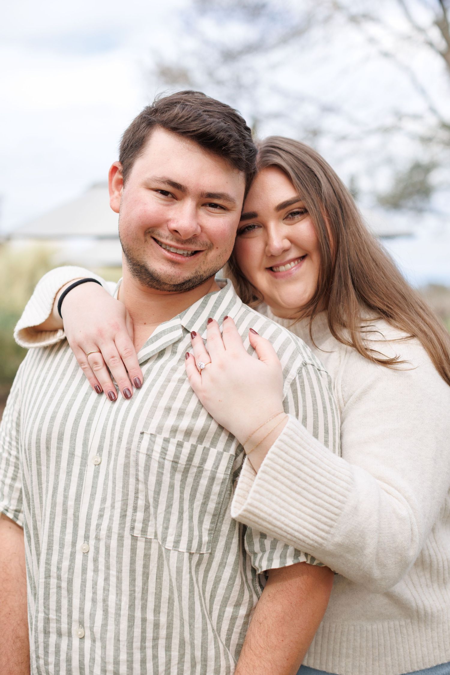 Happy couple embracing outdoors, woman showing engagement ring while smiling at camera.