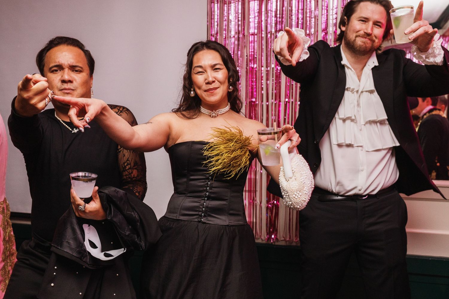 Three friends posing and pointing at a festive party with pink tinsel backdrop, wearing elegant black outfits.