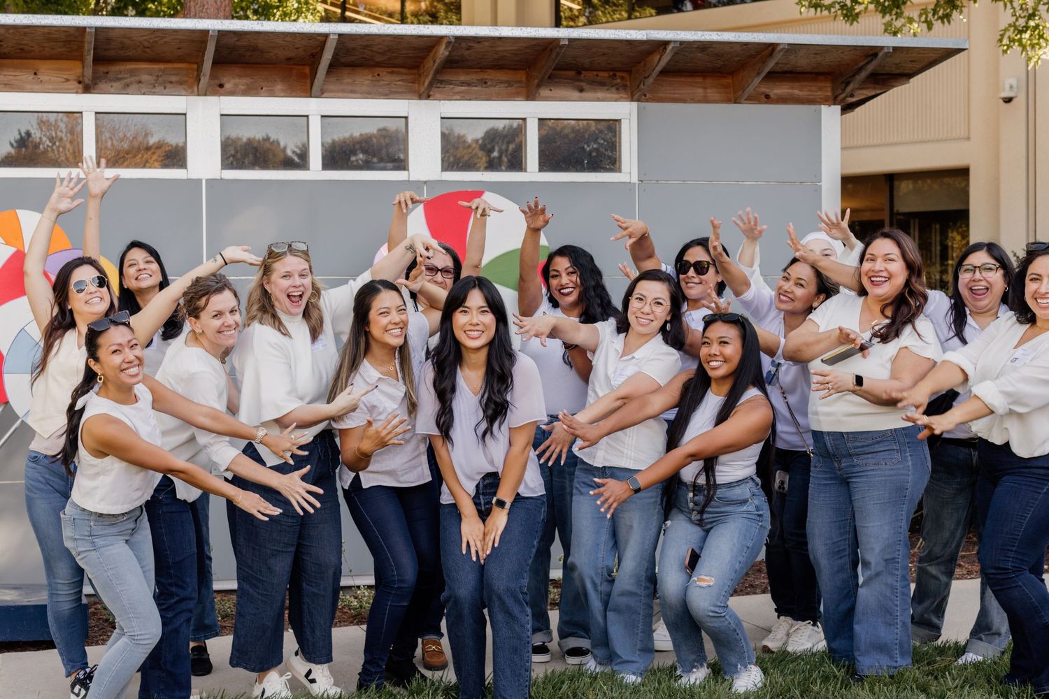 A diverse group of young women in white tops and jeans pose cheerfully outdoors, celebrating together.