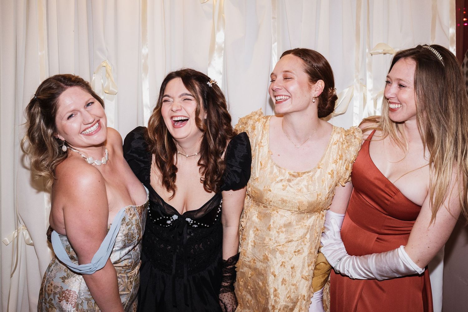 Four women in elegant formal dresses laughing together at a glamorous event.