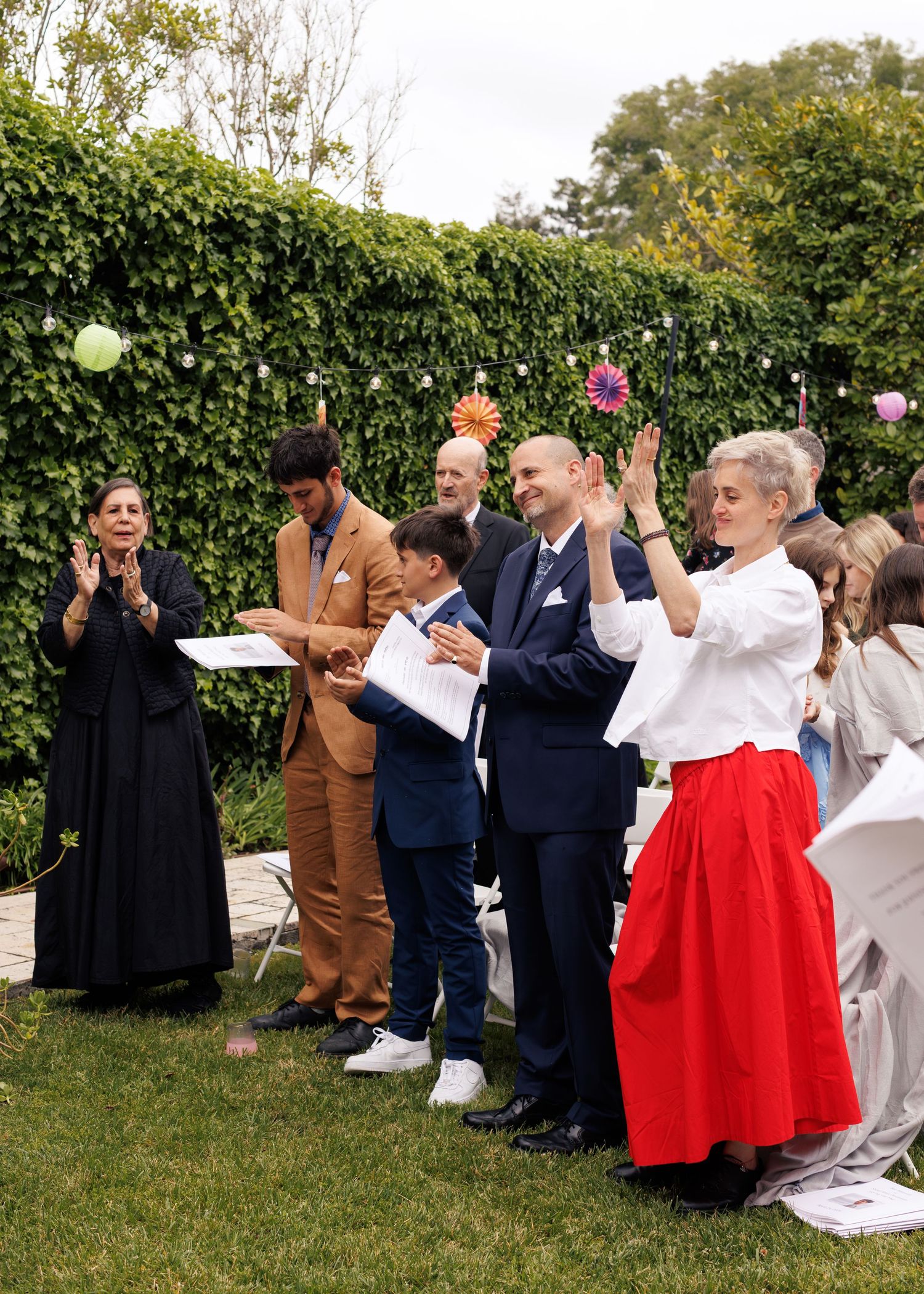 Guests clapping at an outdoor garden wedding ceremony with colorful floral decorations and lush green hedges.