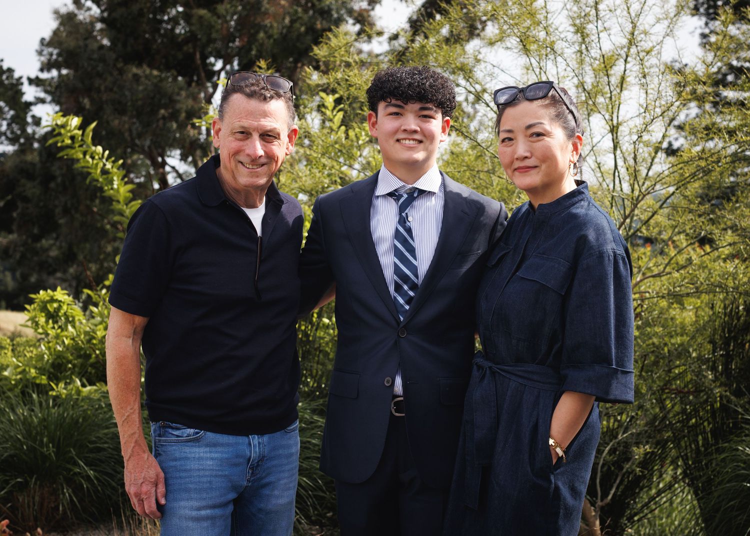 Three people smiling outdoors, young man in suit with striped tie flanked by two adults at a formal celebration.