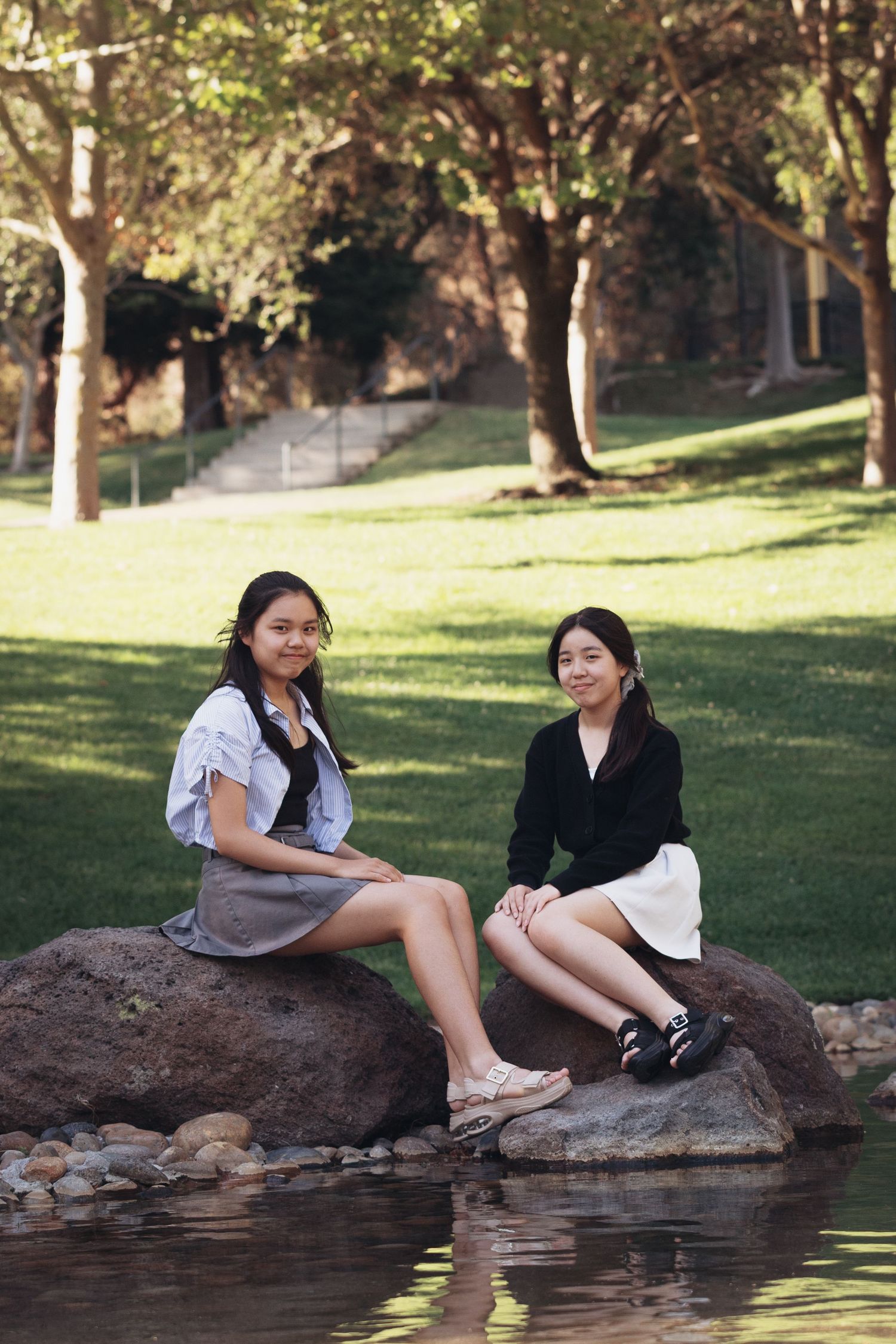 Two young Asian sister sitting on rocks by a pond in a lush green park, during a family photography session in Danville, California.