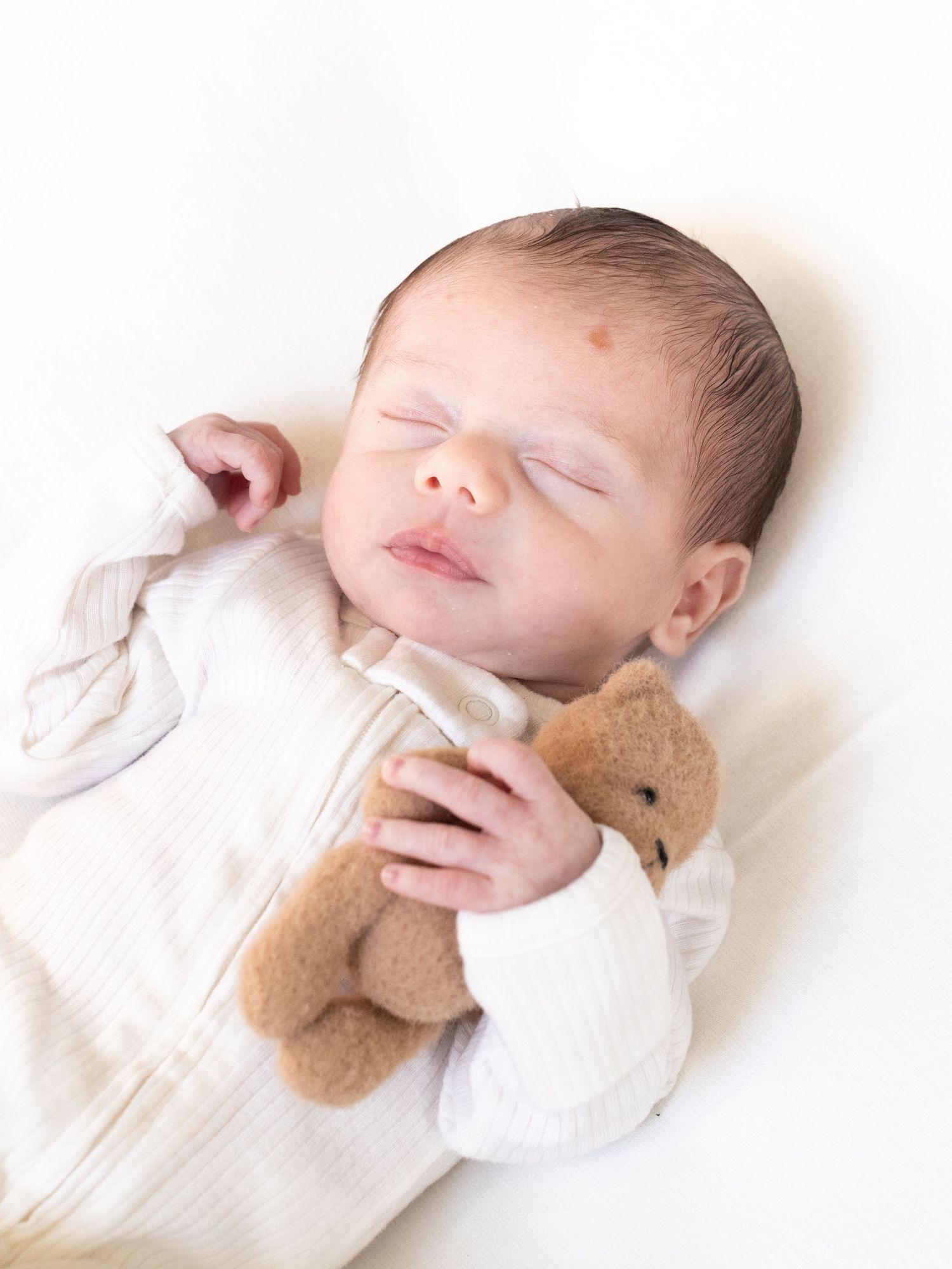 Sleeping newborn baby in white ribbed onesie cuddling a small brown teddy bear on a white background.
