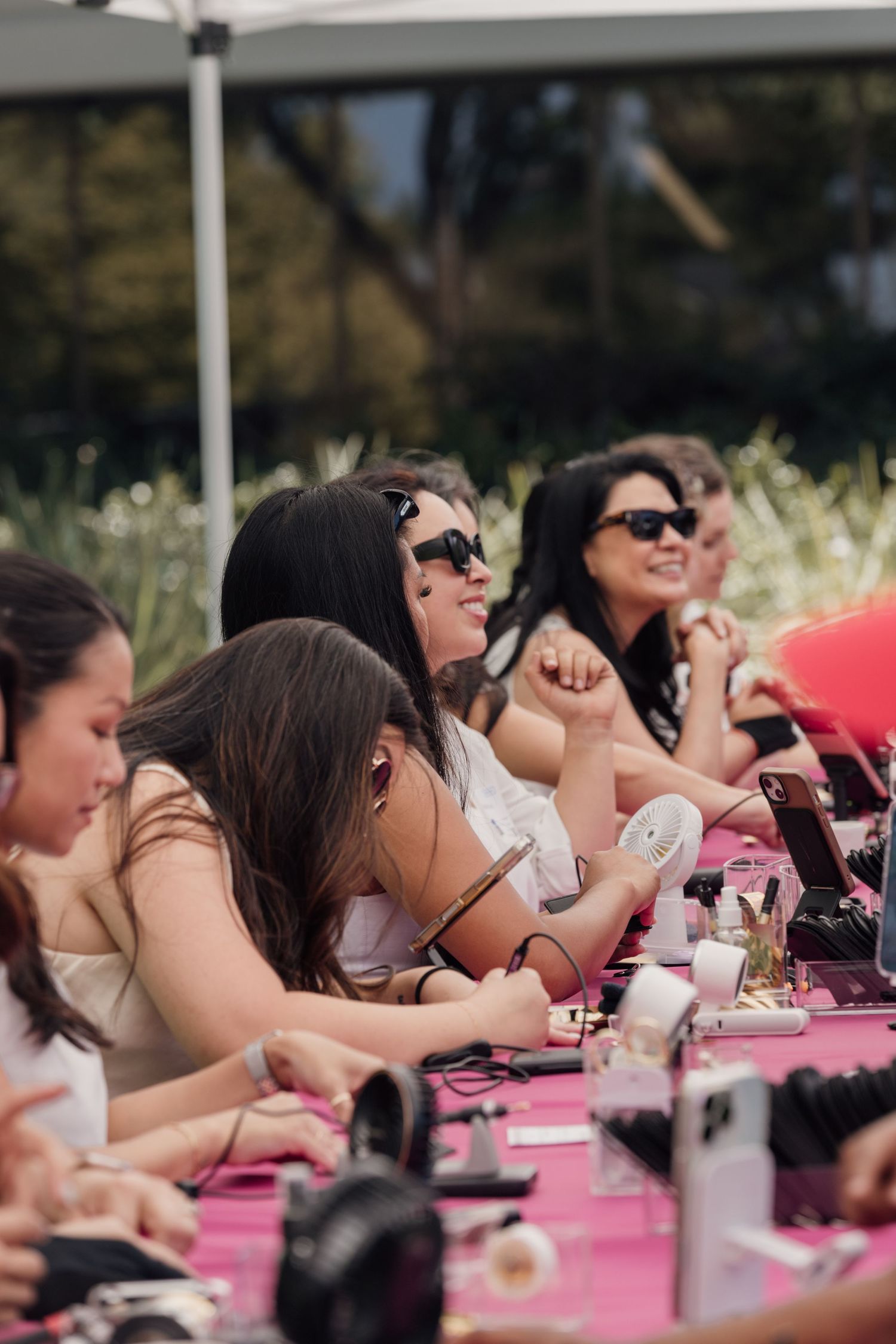 Group of women sitting at a pink table outdoors, smiling and enjoying a beauty or jewelry event.
