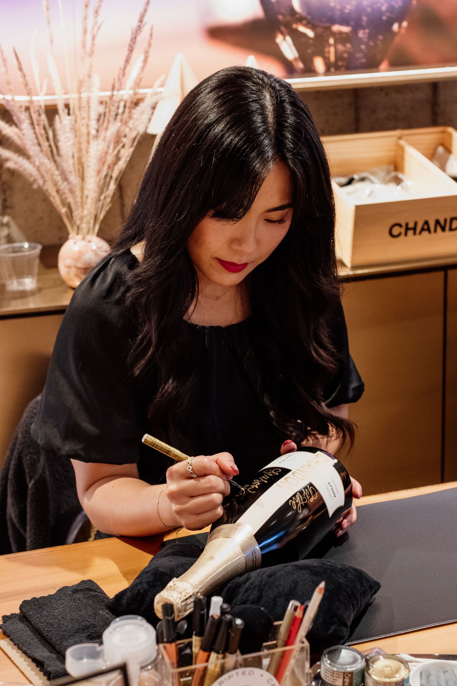 Woman in black dress signing a Chandon champagne bottle with a gold marker at an elegant event.