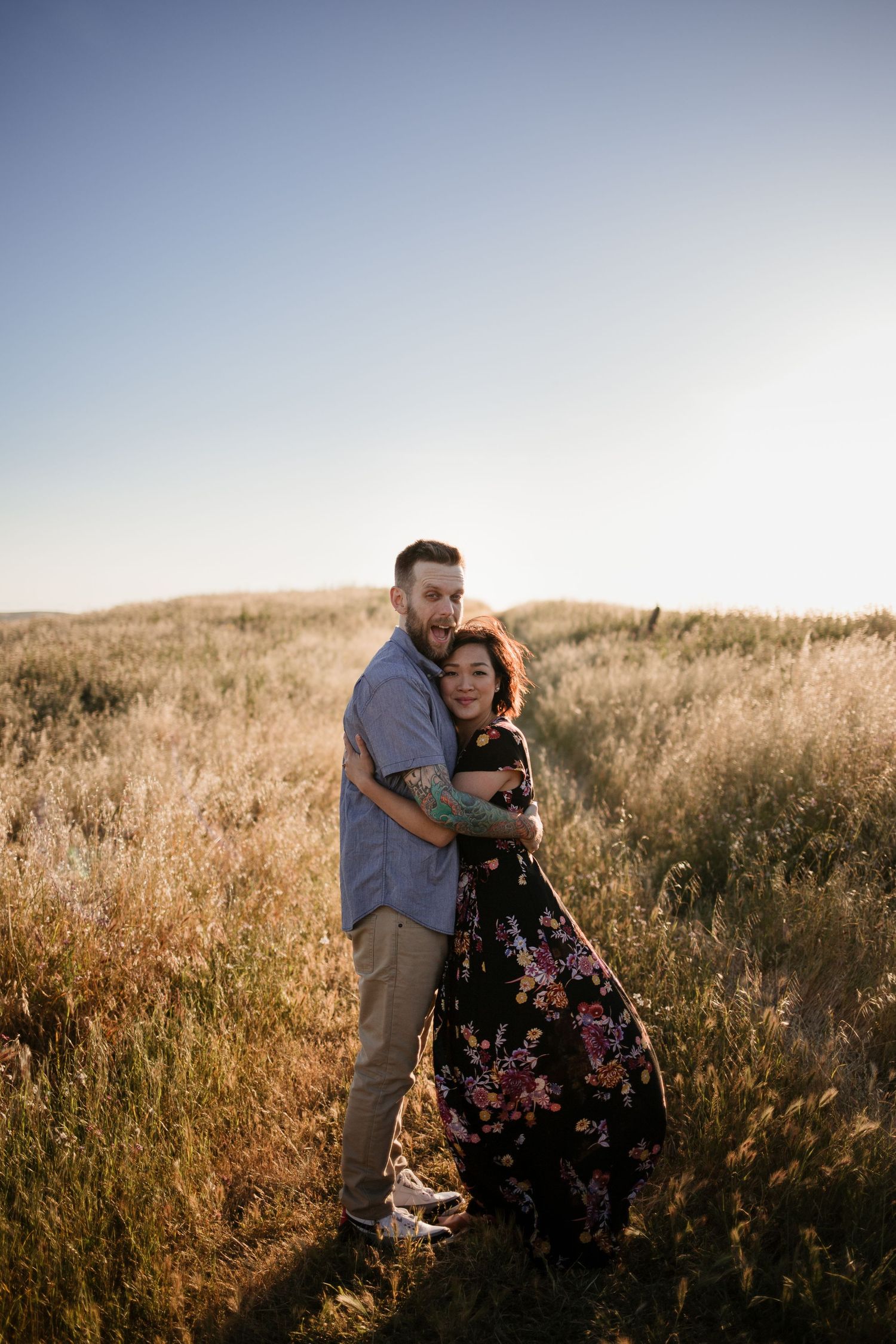 Couple embracing in golden field at sunset, woman in floral dress, man in blue shirt, romantic outdoor portrait.