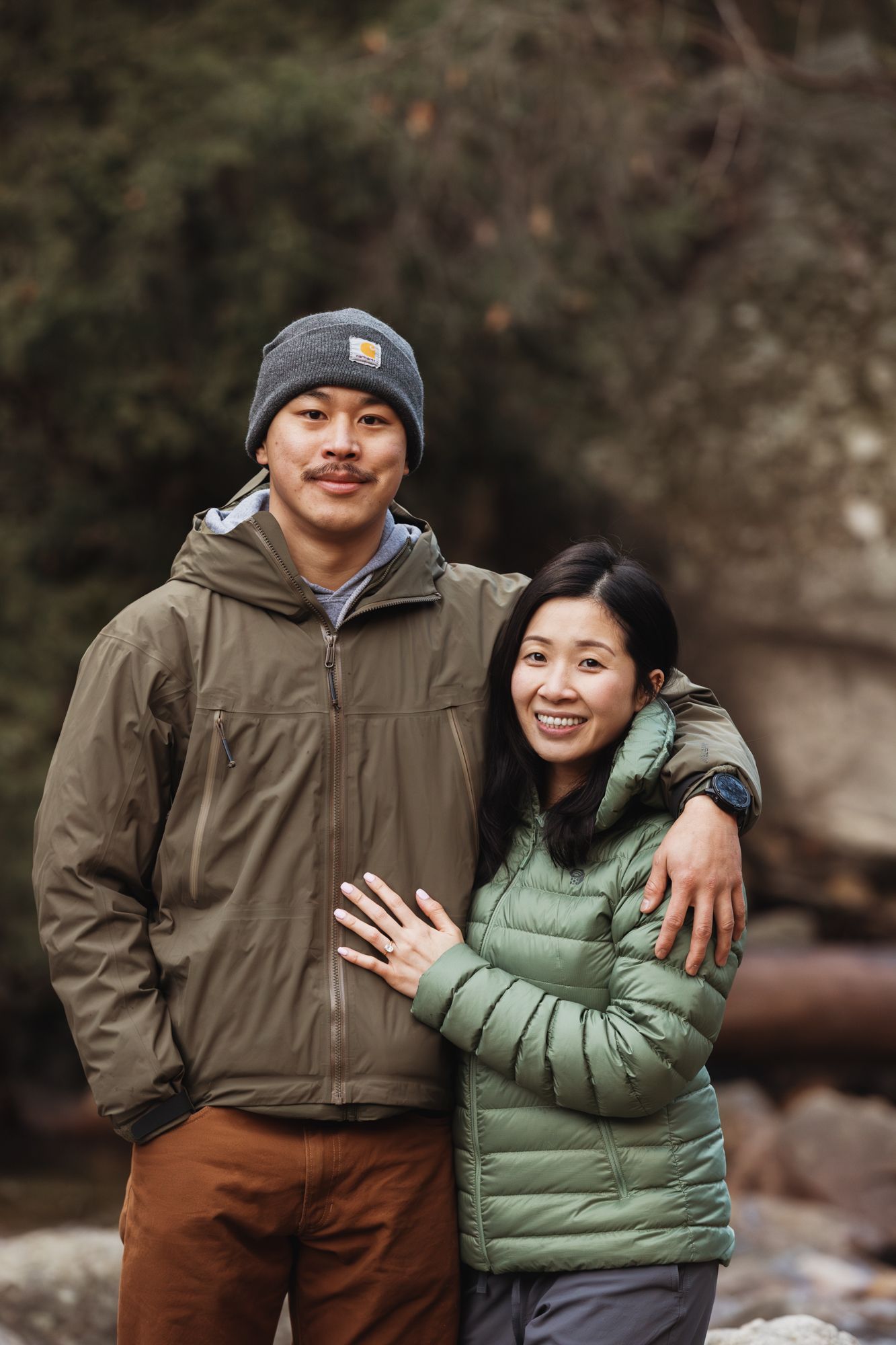 A smiling couple in green jackets poses outdoors near a rocky stream, the woman displaying an engagement ring.