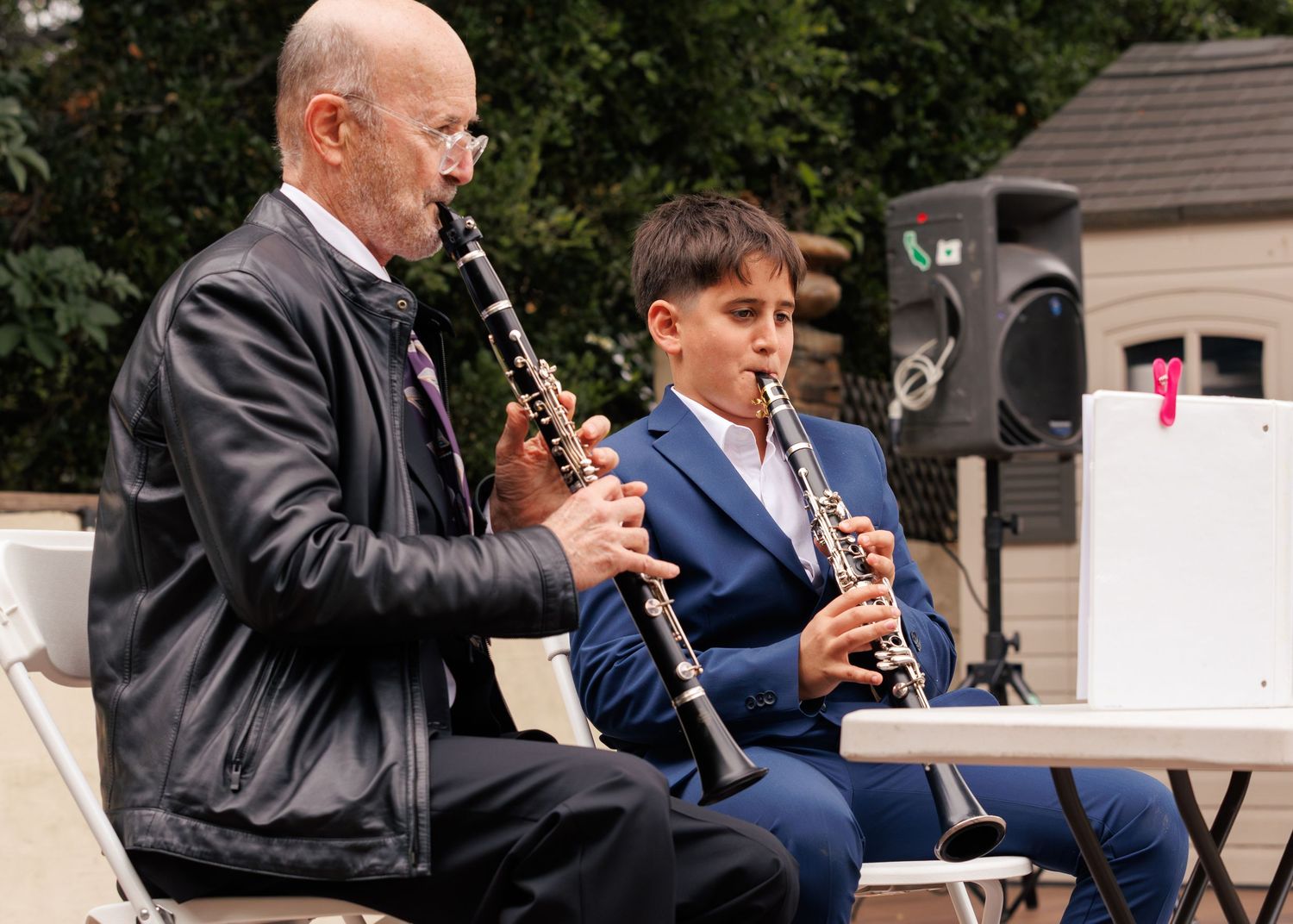 An older man and a young boy in a blue suit play clarinets together outdoors at a music performance.