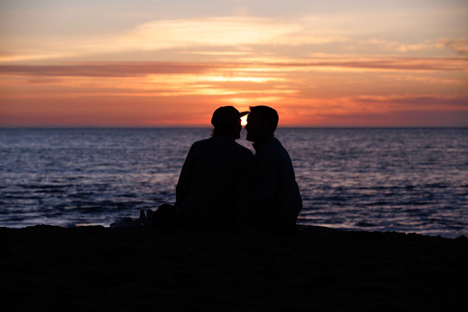 Silhouette of couple sitting together on beach shore watching vibrant orange and red sunset over ocean horizon.