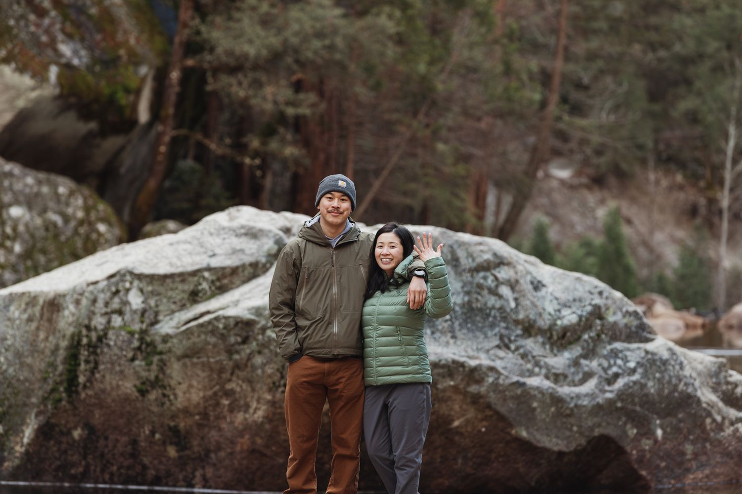 Couple smiling and posing in front of large rocks in a forest during a winter hike.