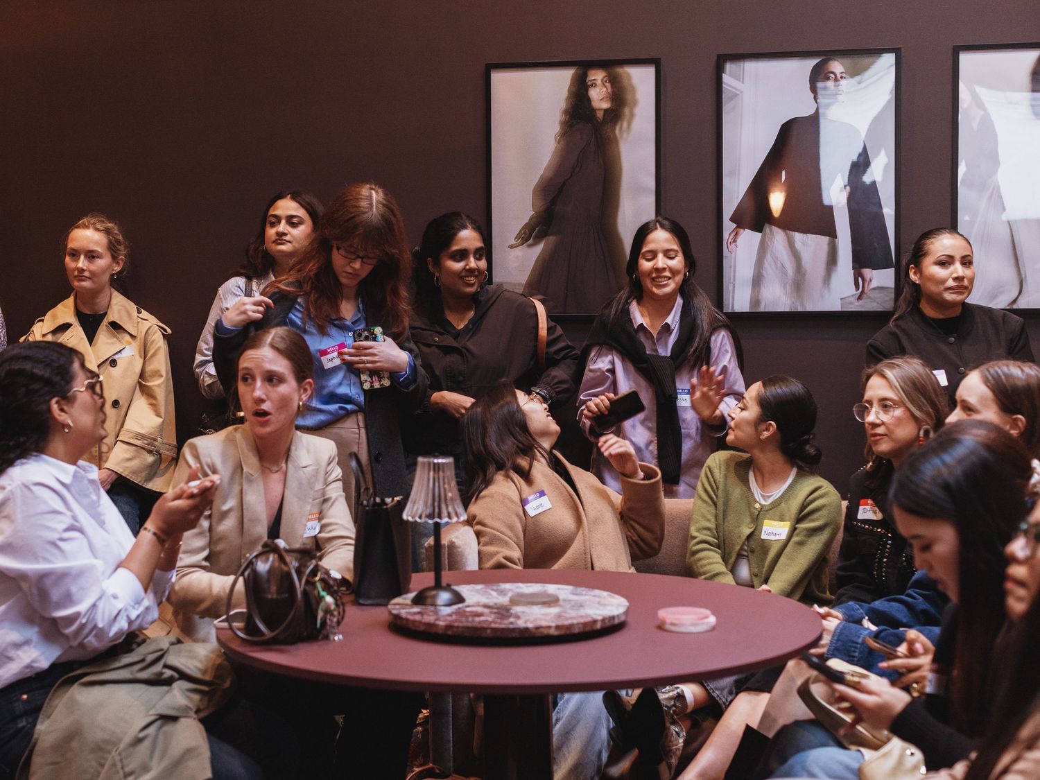 A diverse group of women gathered around a round table at a stylish indoor event, socializing and holding drinks.